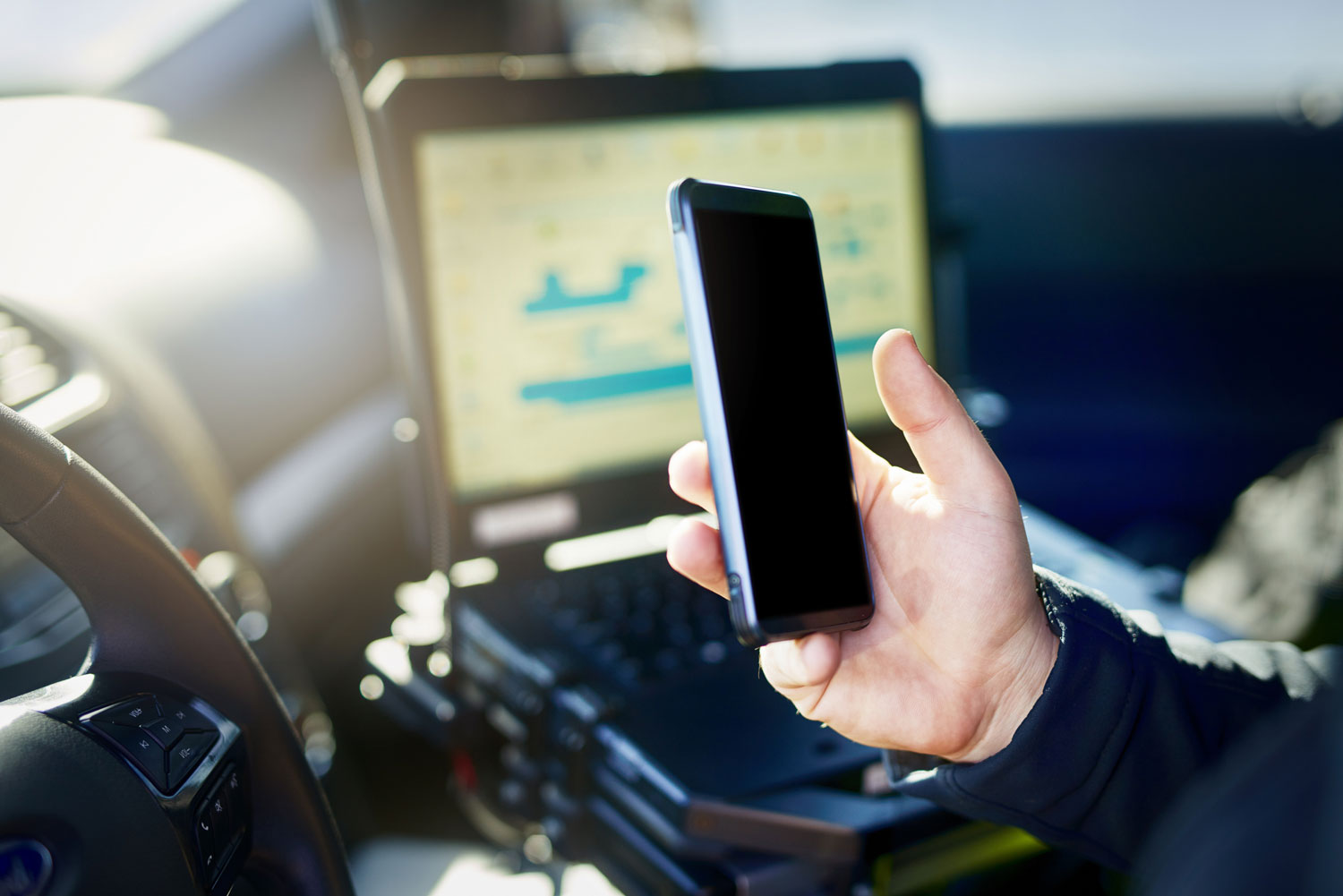 Officer holding a smartphone beside a mounted computer inside a patrol vehicle